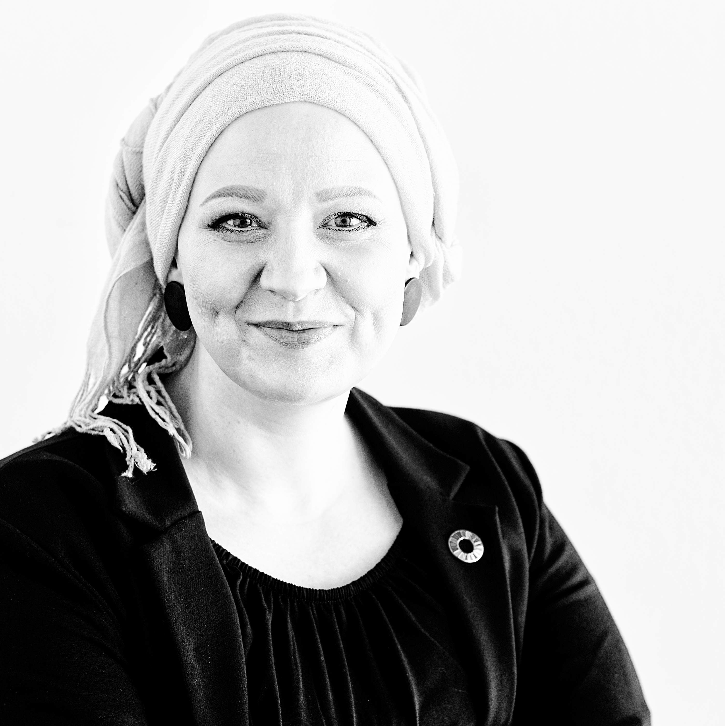 Das Bild zeigt eine lächelnde Frau mit hellem Kopftuch, in dunklem Oberteil und Blazer mit einem Pin der Sustainable Development Goals auf dem Revers.