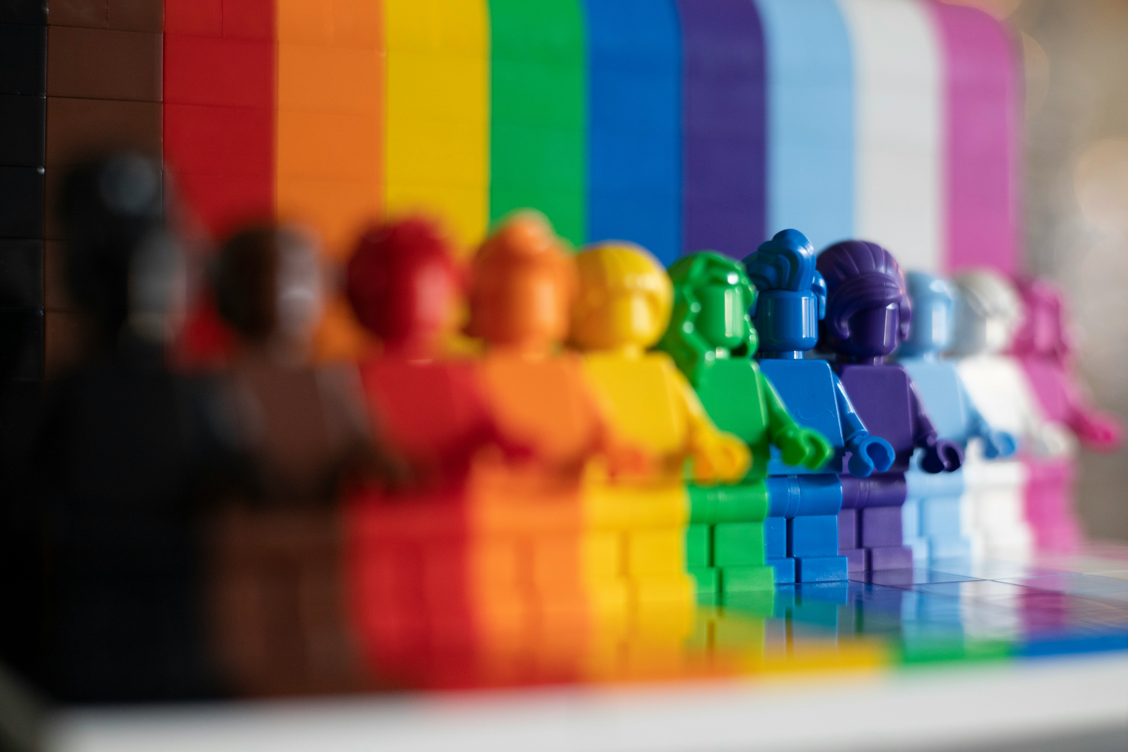 Das Bild zeigt eine eine Reihe bunter Legofiguren, die vor einem farbenfrohen, prismatischen Hintergrund stehen, wobei die Gesichter unscharf sind und die Farben des Hintergrunds dominieren.