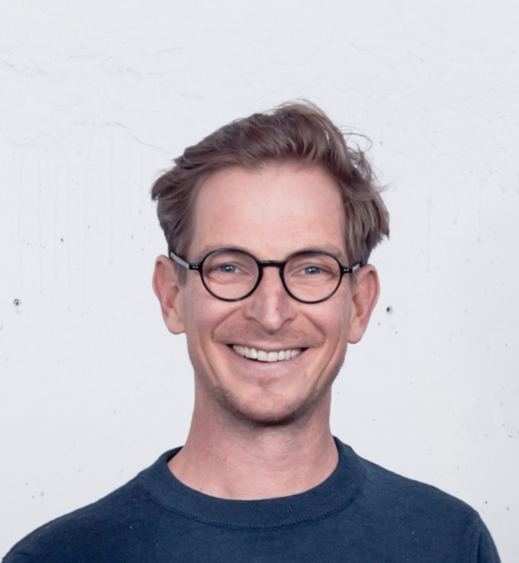 KI generiert: Das Bild zeigt einen lächelnden Mann mit kurzen, dunklen Haaren und einer Brille, der ein dunkelblaues T-Shirt trägt.