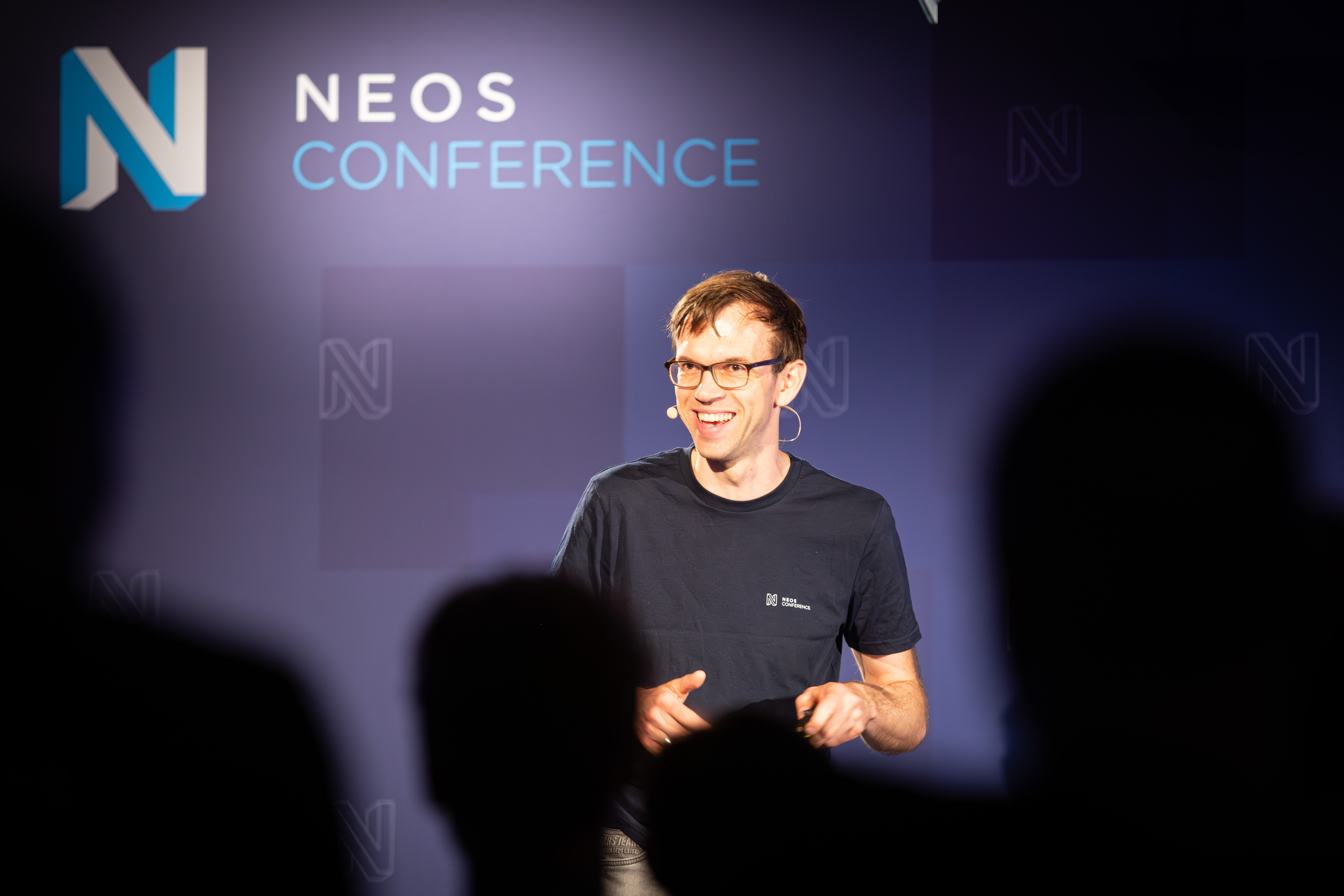 KI generiert: Das Bild zeigt einen lächelnden Mann in einem grauen T-Shirt, der vor einem Hintergrund mit dem Aufdruck "NEOS CONFERENCE" steht und eine Armbanduhr trägt.