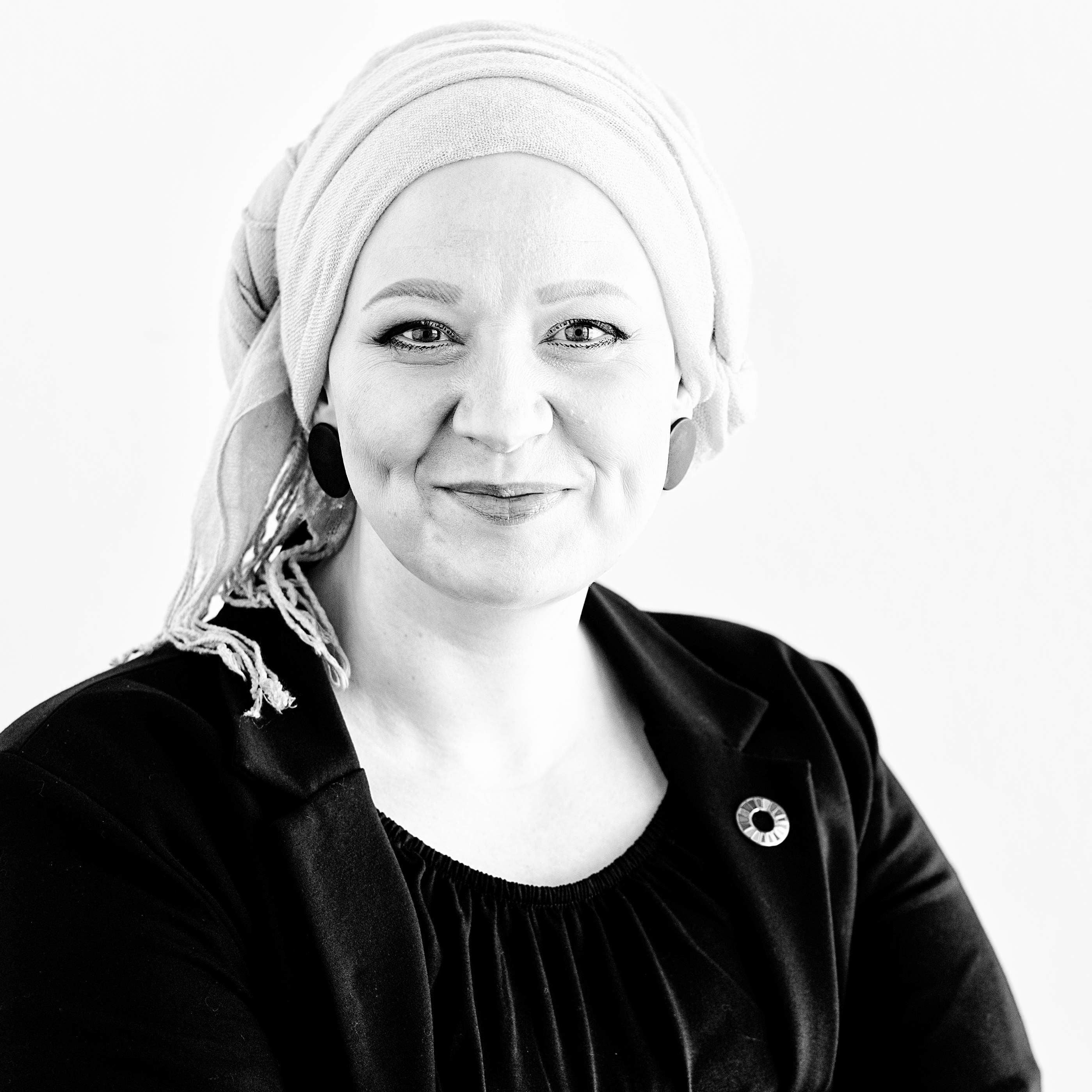 Das Bild zeigt eine lächelnde Frau mit hellem Kopftuch, in dunklem Oberteil und Blazer mit einem Pin der Sustainable Development Goals auf dem Revers.