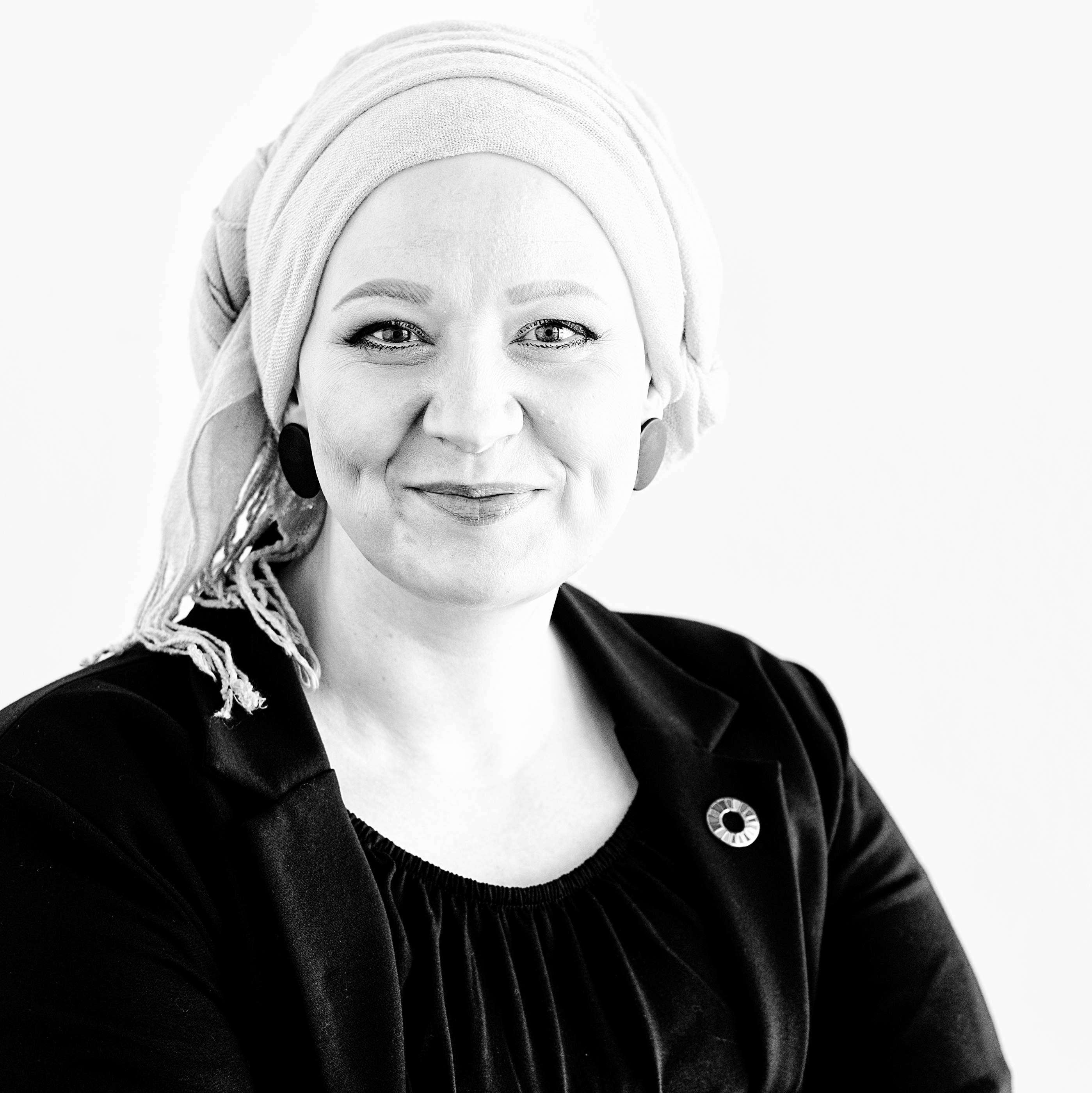 Das Bild zeigt eine lächelnde Frau mit hellem Kopftuch, in dunklem Oberteil und Blazer mit einem Pin der Sustainable Development Goals auf dem Revers.