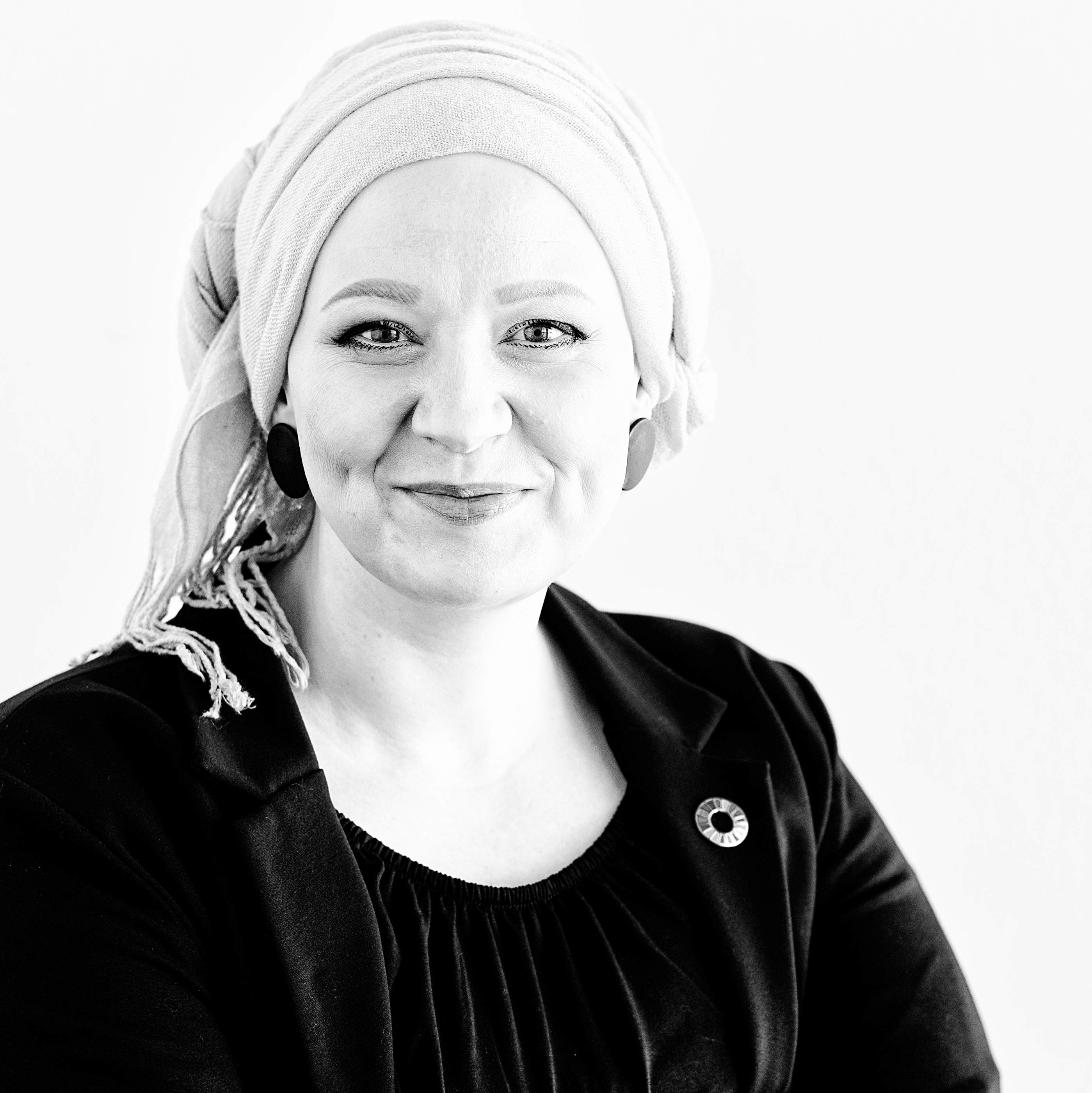 Das Bild zeigt eine lächelnde Frau mit hellem Kopftuch, in dunklem Oberteil und Blazer mit einem Pin der Sustainable Development Goals auf dem Revers.
