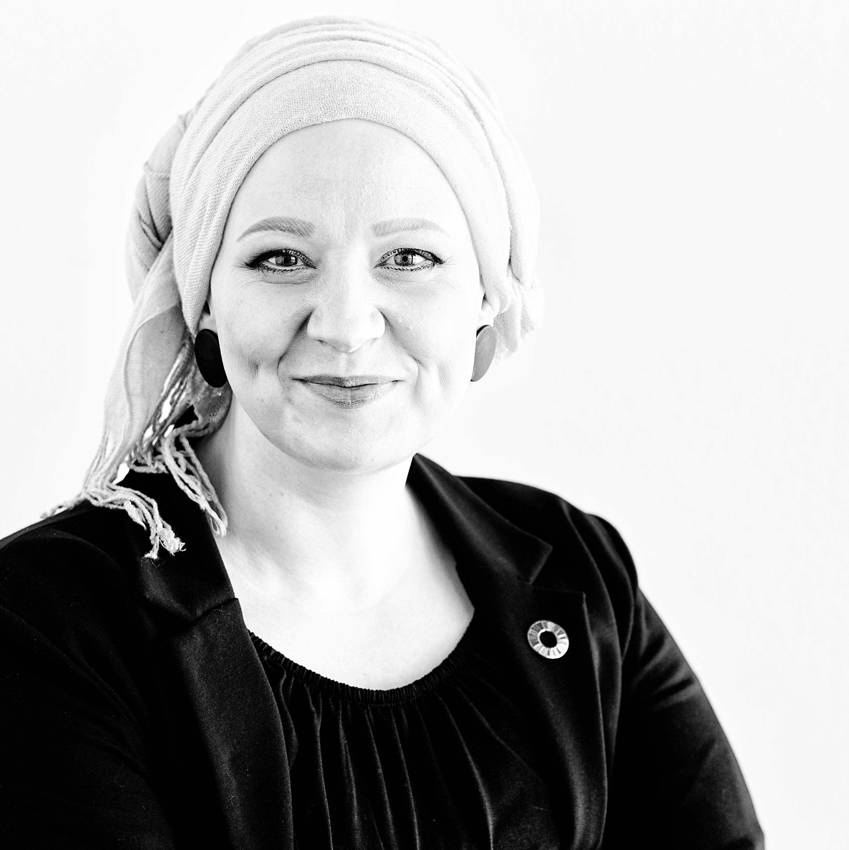 Das Bild zeigt eine lächelnde Frau mit hellem Kopftuch, die ein schwarzes Oberteil und einen schwarzen Blazer mit einem Button der Sustainable Development Goals trägt.